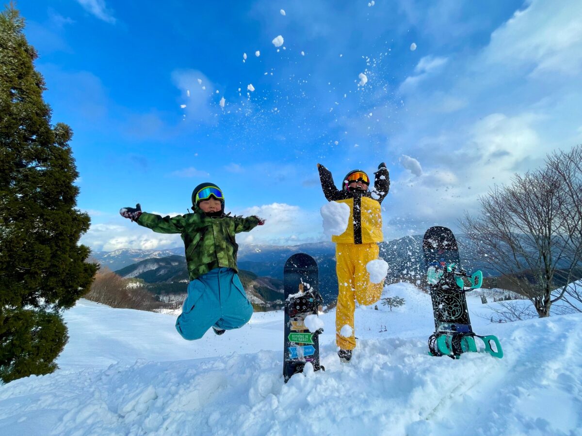 青空の下で雪をまき上げてジャンプする親子のスノーボーダー